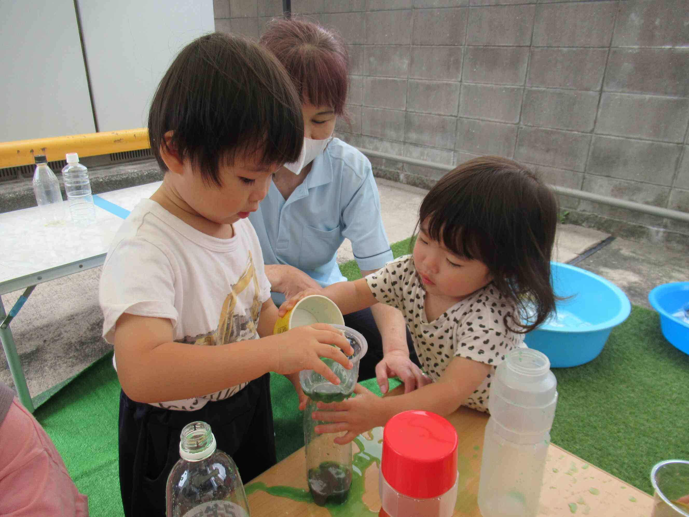「持っててあげるね」ペットボトルに水を移し替える2歳児Áくんの様子を見て手伝いを始める1歳児Rちゃん。異年齢での関わりも自然とみられます。